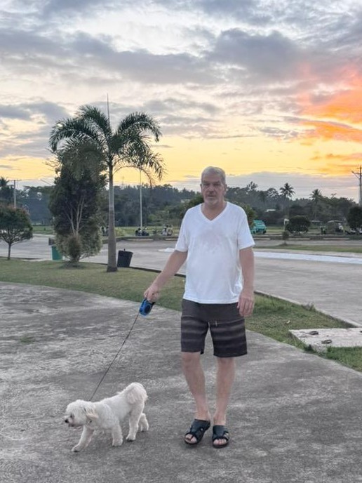 Man in his 60s walking a white dog at sunset along a tropical boulevard — active recovery through light movement and social connection as part of a longevity lifestyle.