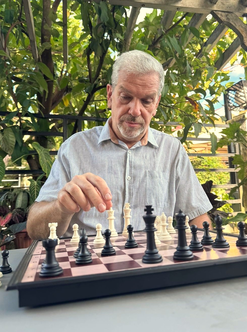 Man in his 60s playing chess outdoors under a pergola surrounded by tropical plants — practicing cognitive training for longevity and mental performance after 40.