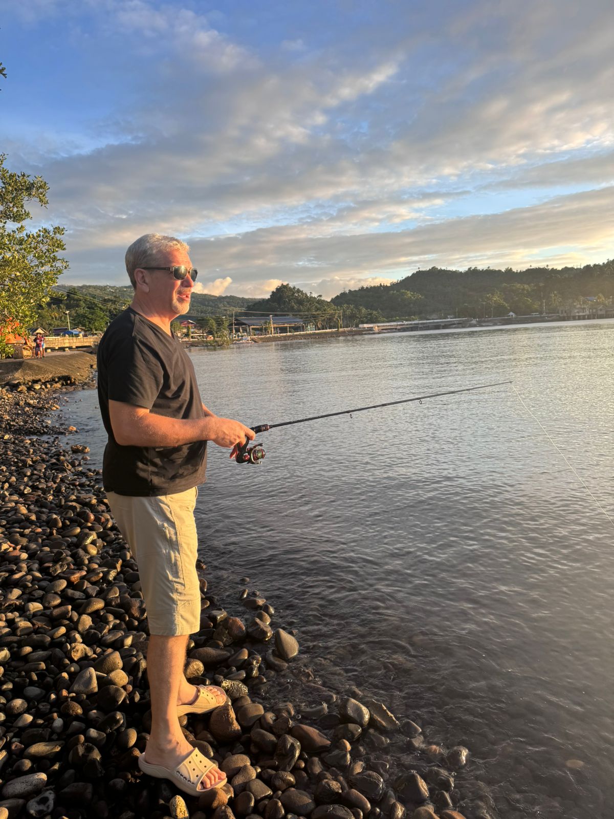 Man in his 60s fishing at golden hour on a rocky shoreline — active recovery through nature and mental rest as part of a longevity training system after 40.